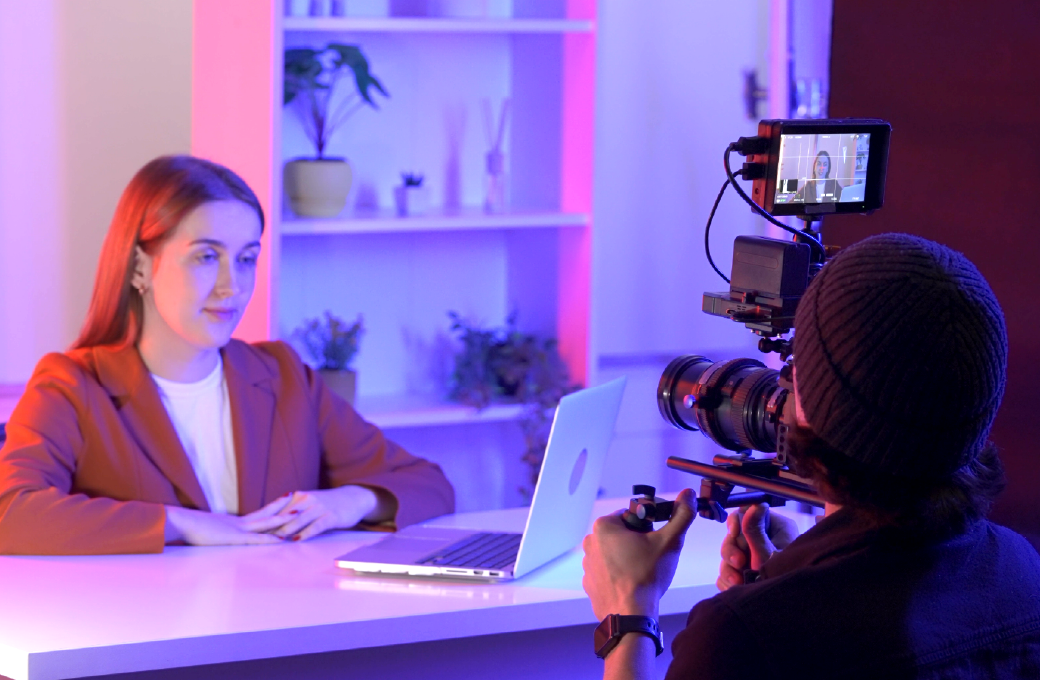 A woman sits at a desk with a laptop while a person films her using a professional camera; shelves with plants are in the background.